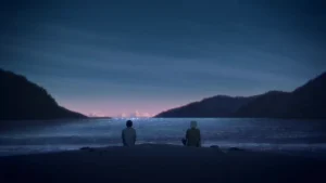 Two people sit side by side on a beach at dusk, gazing out over a calm, shimmering sea. The sky is painted in deep blues and purples, with hints of pink light on the horizon. Hills rise on either side of the bay, framing the serene, contemplative scene.