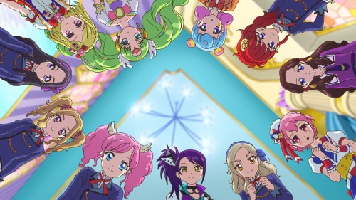 “Aikatsu! × PriPara THE MOVIE” – Collaboration Stage Footage and New Stills Unveiled 1 Aikatsu! movie scene: Twelve colorful idol girls lean in a full circle toward the camera from above, forming a bright ring of uniforms, bows, twin‑tails, curls, and vivid hair colors around a radiant skylight ceiling with a starburst pattern. The low, upward perspective frames their eager, supportive expressions before a performance or pact.