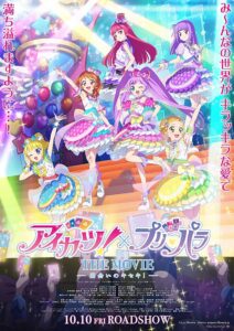 Aikatsu! × PriPara The Movie crossover poster: Five glittering idol girls strike energetic poses on a rainbow‑lit stage—frilly layered skirts, candy‑colored ribbons, star and heart motifs—against a backdrop of balloons, floating glowsticks, and a giant spinning centerpiece crowned with lights. Audience silhouettes at the bottom hold penlights that shower sparkling beams upward. Bold joint franchise logo with THE MOVIE subtitle spans the center above a 10.10 roadshow date.