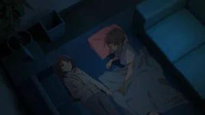 Nighttime anime scene viewed from above: two teens lie on a blue floor beside a low sofa—one girl in a light hoodie and jeans gazing upward, the other (a boy) under a thin gray blanket with a pink pillow, turned toward her as they talk quietly in the dim, cool-toned room. A plant, trash bin, and low TV stand sit nearby in shadow.s